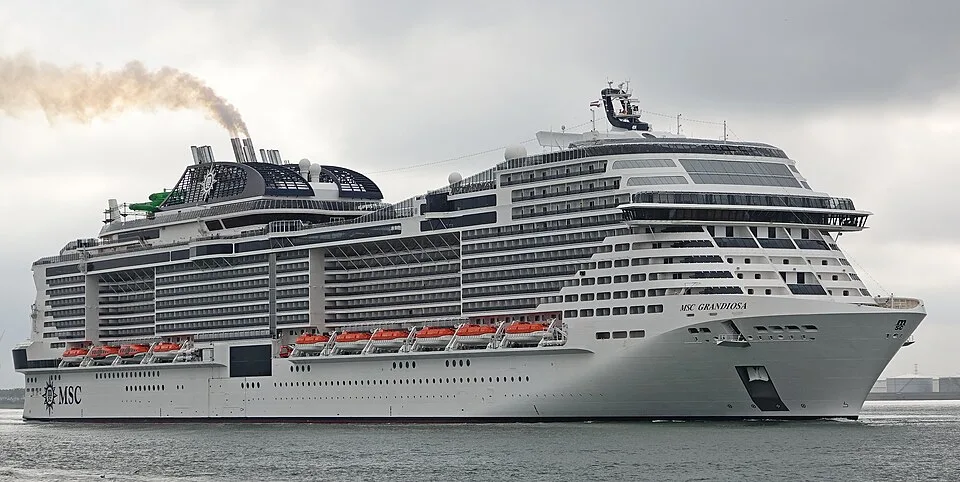 The massive cruise ship MSC Grandiosa sails on open water under an overcast sky. The white vessel features a tiered front, numerous cabin balconies, and bright orange lifeboats along the side. Dark exhaust smoke rises from its large black funnel.
