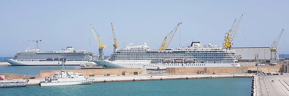 Two large, white cruise ships are docked at a harbor terminal surrounded by several tall yellow construction cranes. A smaller naval or security boat is moored in the foreground water next to a concrete pier under a clear, bright sky.