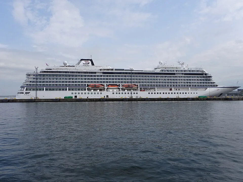 A large white Viking cruise ship docked along a long flat pier under a cloudy sky. The multi-tiered ship features rows of windows, stateroom balconies, and several bright orange lifeboats hanging along the center of its hull.