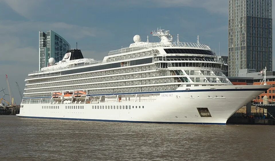 A large white cruise ship named "Viking Sky" docked along a city waterfront. The modern vessel features tiered decks with glass balconies, orange lifeboats tucked on the side, and a black smokestack, with tall skyscrapers visible in the background under a hazy sky.