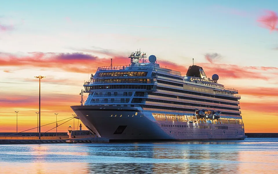 The large Viking Sky cruise ship sits docked in calm harbor water at sunset. Its multiple decks and indoor spaces glow with warm golden lights. The background features a dramatic, vibrant sky filled with orange, pink, and blue clouds, which cast colorful reflections across the water.