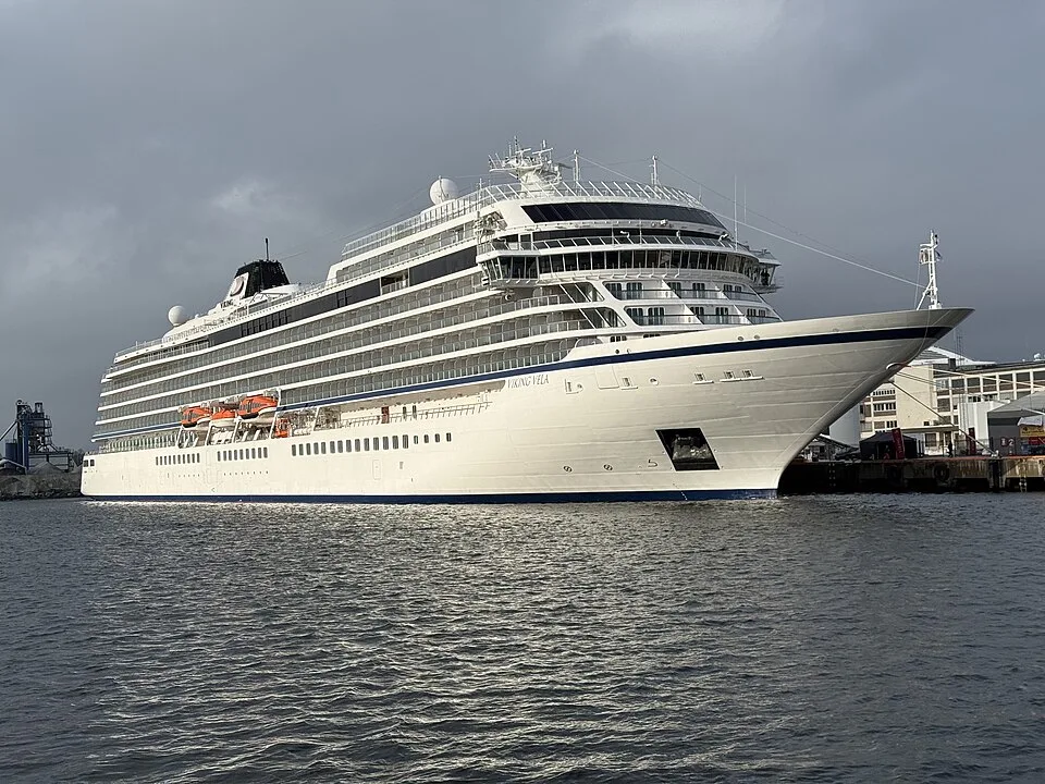 A large white cruise ship named "Viking Vela" docked in a harbor on choppy grey water. The multi-deck vessel features glass balcony railings, a black smokestack, and orange lifeboats along the side, set against an industrial shoreline under a dark, overcast sky.