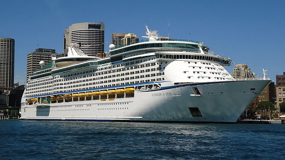 A large white cruise ship named "Voyager of the Seas" docked in a harbor on deep blue water. The ship shows multiple decks, yellow lifeboats, and distinctive round windows on its bow, with a cityscape of tall skyscrapers under a clear blue sky in the background.