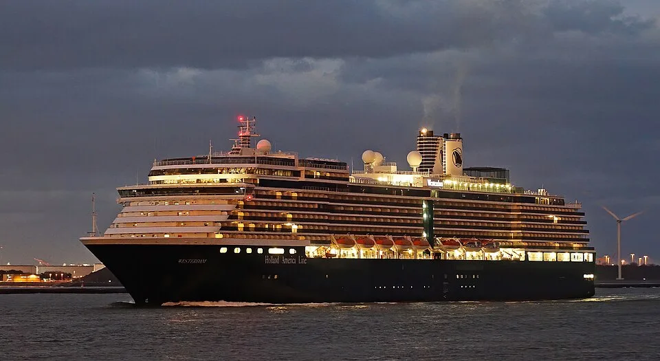 The Holland America Line cruise ship MS Westerdam sails at dusk with its decks brightly illuminated. The ship features a dark hull, multiple tiers of lit windows, and white smokestacks. A wind turbine and distant port lights are visible under a cloudy twilight sky.