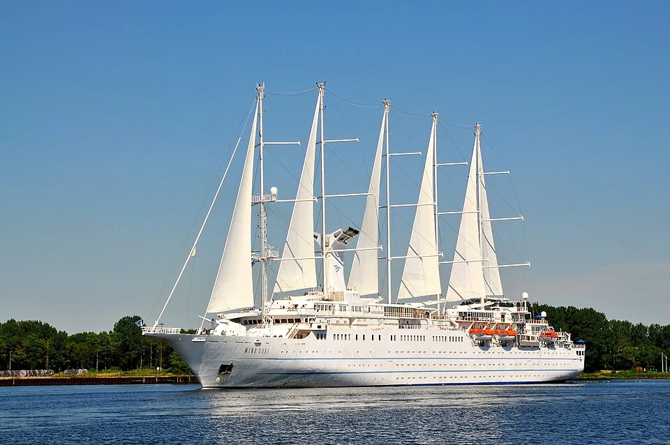 A large white sailing cruise ship with multiple tall masts and billowing sails traveling across calm blue water under a clear sky, with a tree-lined shoreline in the background.
