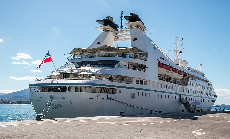 A white cruise ship is docked at a pier under a clear blue sky. It has two distinctive funnel towers, a canopy deck, and rows of small windows. A flag flies from the stern, where "Star Breeze" is faintly visible. The ship is secured to the concrete dock with heavy mooring lines.