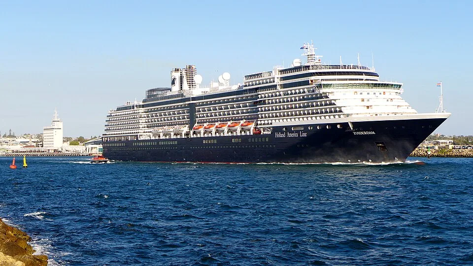 The Holland America Line cruise ship Zuiderdam cruising on deep blue water. The massive vessel features a dark hull, stacked white passenger decks, a row of orange lifeboats, and a white funnel under a clear sky.