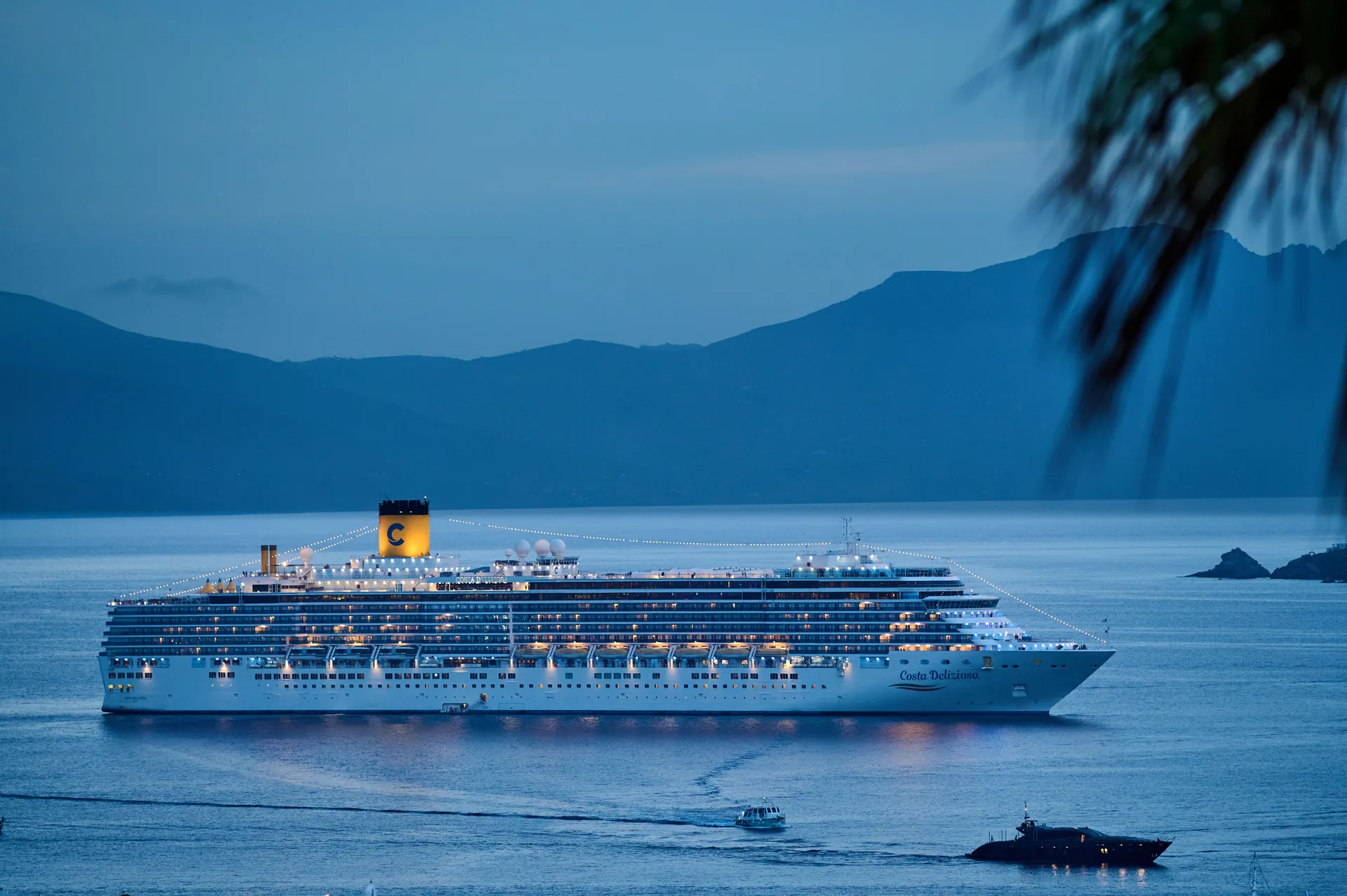 The Costa Deliziosa lit up at dusk, lying at anchor in a bay below dark hills